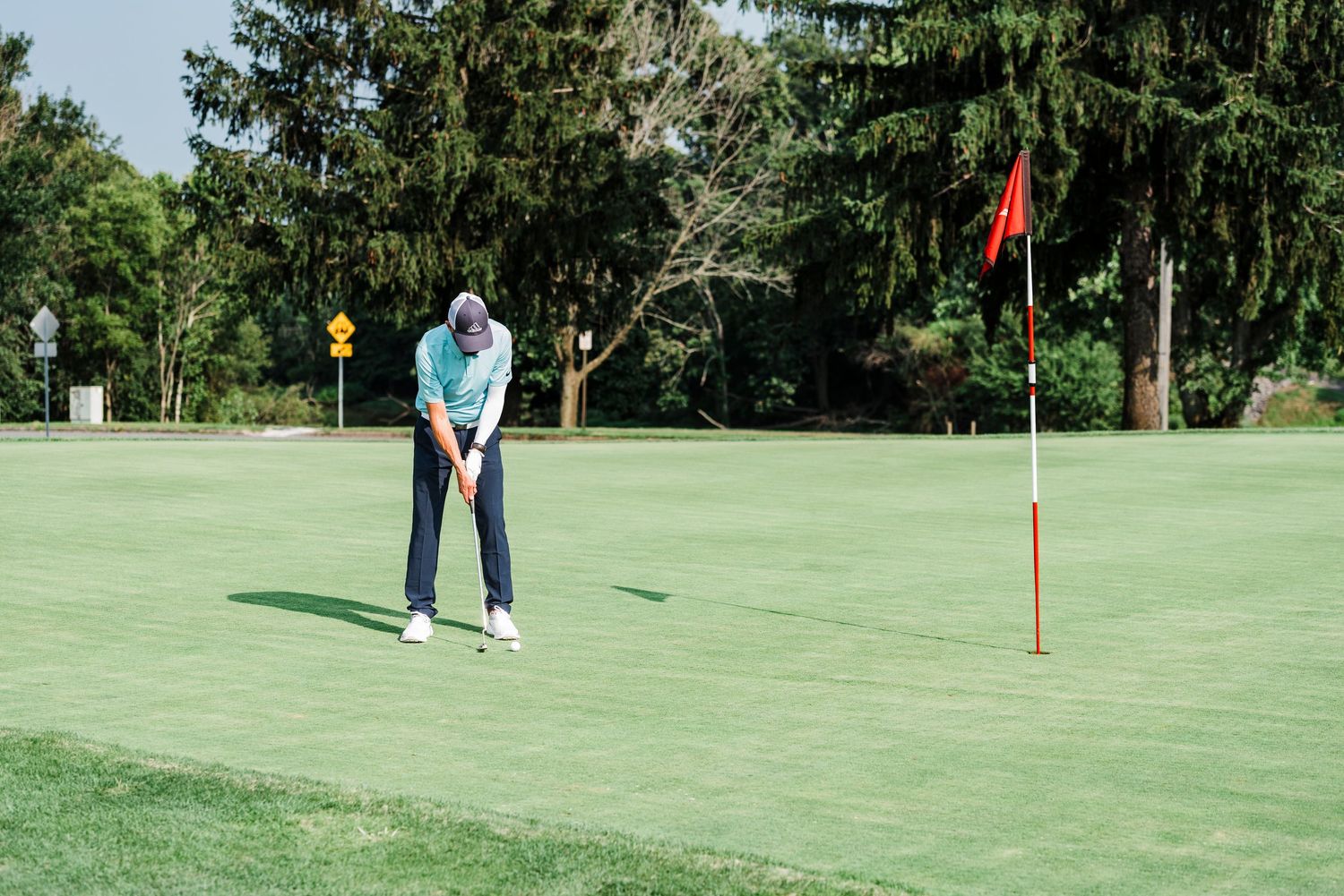 Golfer in light blue shirt lines up putt on manicured green course with red flag pin visible.