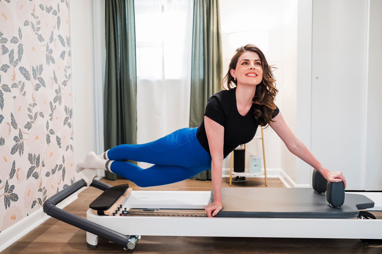 Person exercising on a reformer Pilates machine in a bright room with floral wallpaper and green curtains.