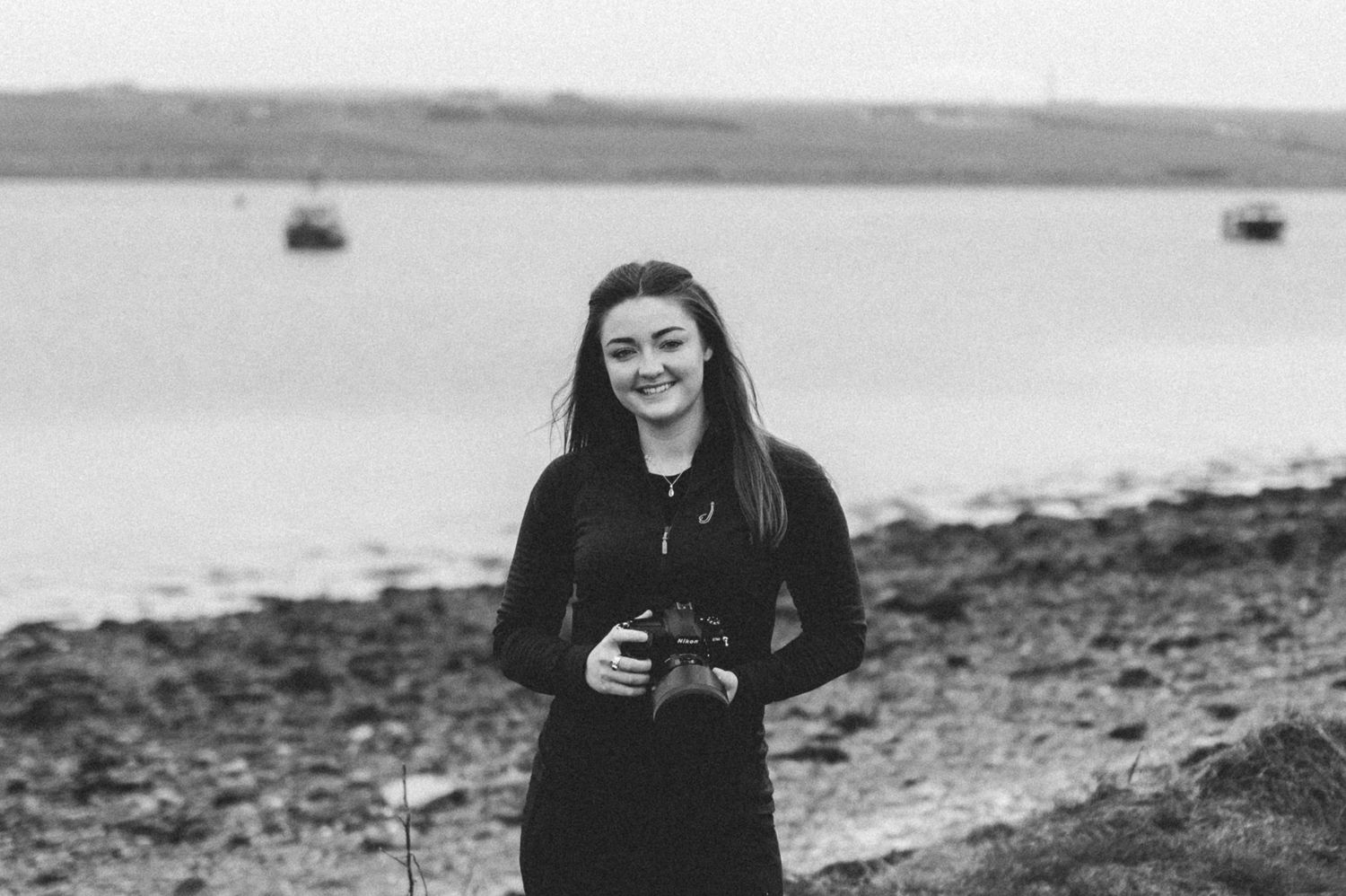 A photographer in a black sweater stands on a rocky shoreline holding a camera with water and boats in the background.