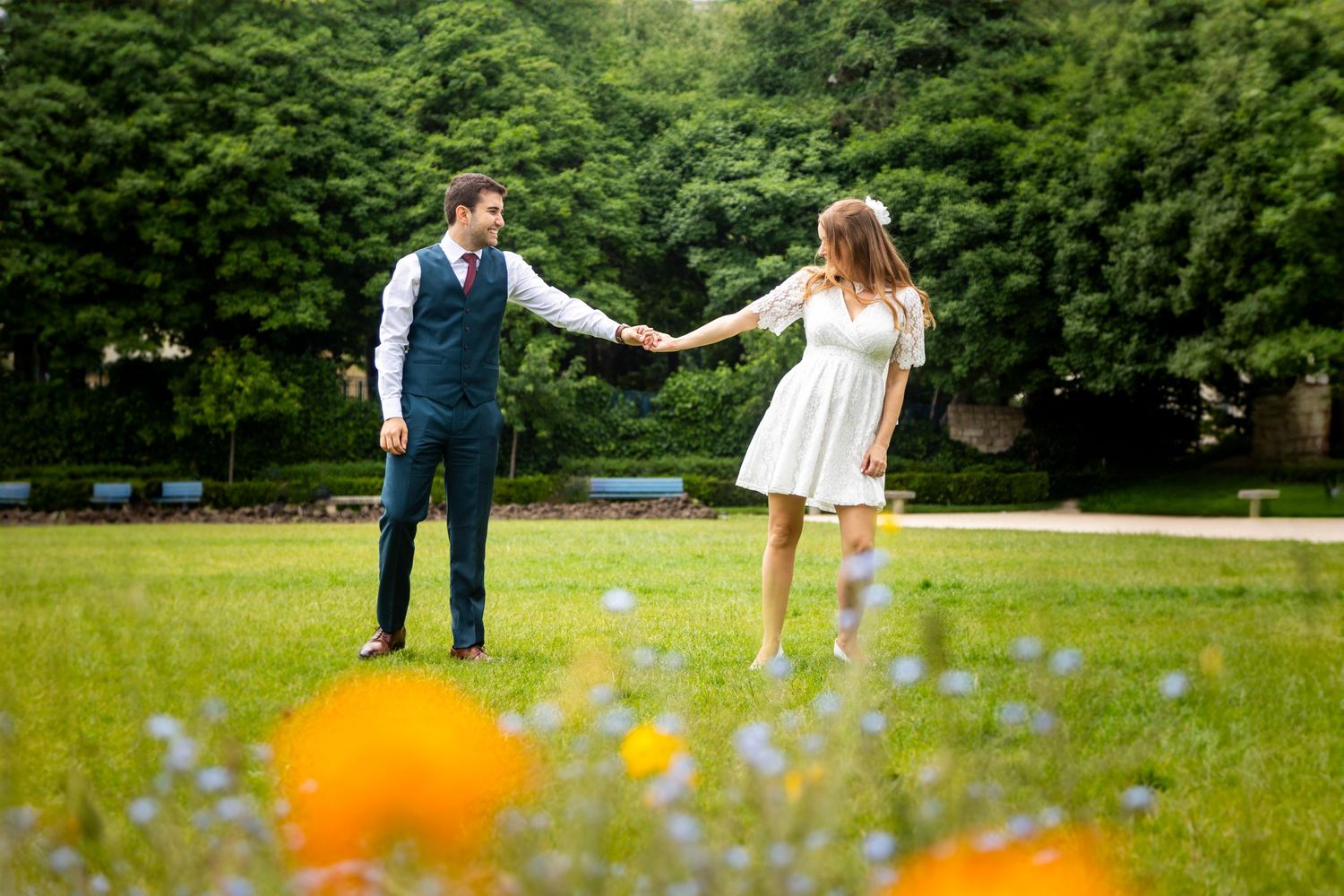 A couple holds hands while dancing in a summer meadow with colorful orange wildflowers in the foreground.