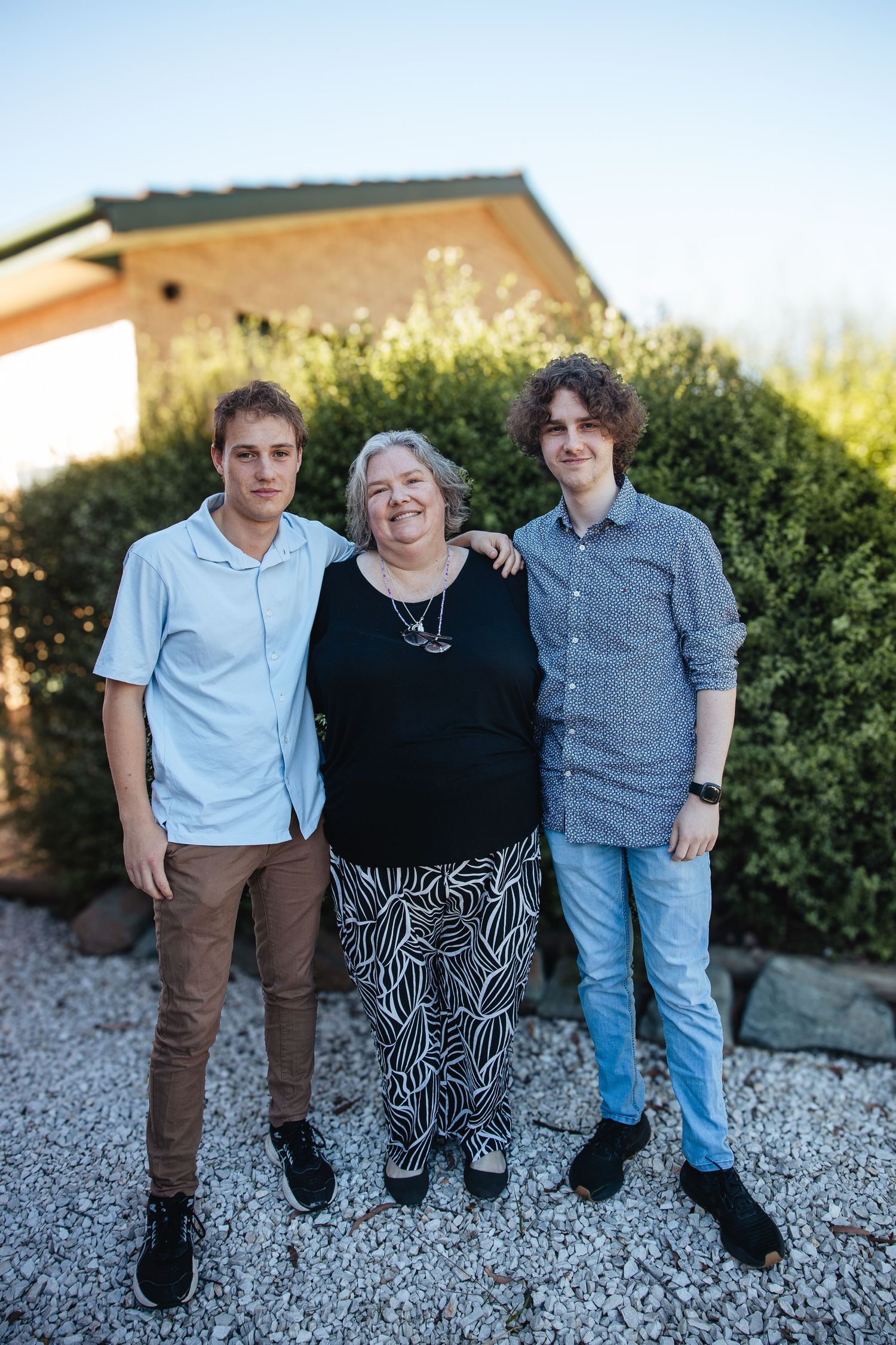 Three people stand together on gravel in front of green shrubs and a brick building on a sunny day.