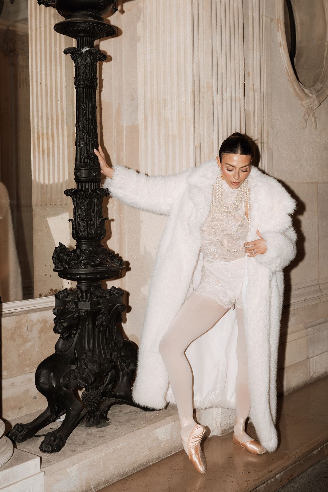A ballet dancer in an all-white ensemble with fur coat poses elegantly against an ornate black pillar.