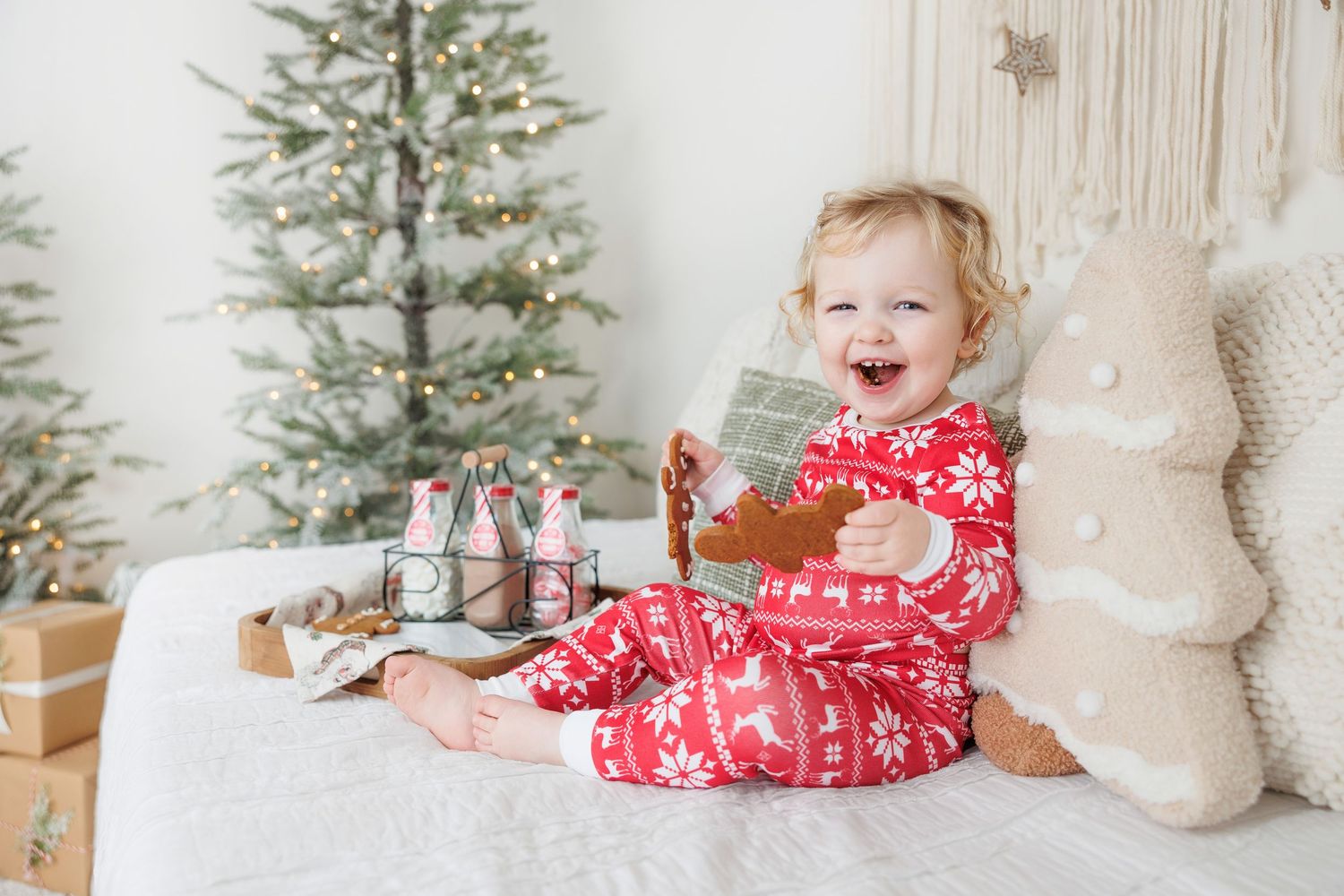 A toddler in red holiday pajamas sits on a white bed with a Christmas tree and plush bear in the background.