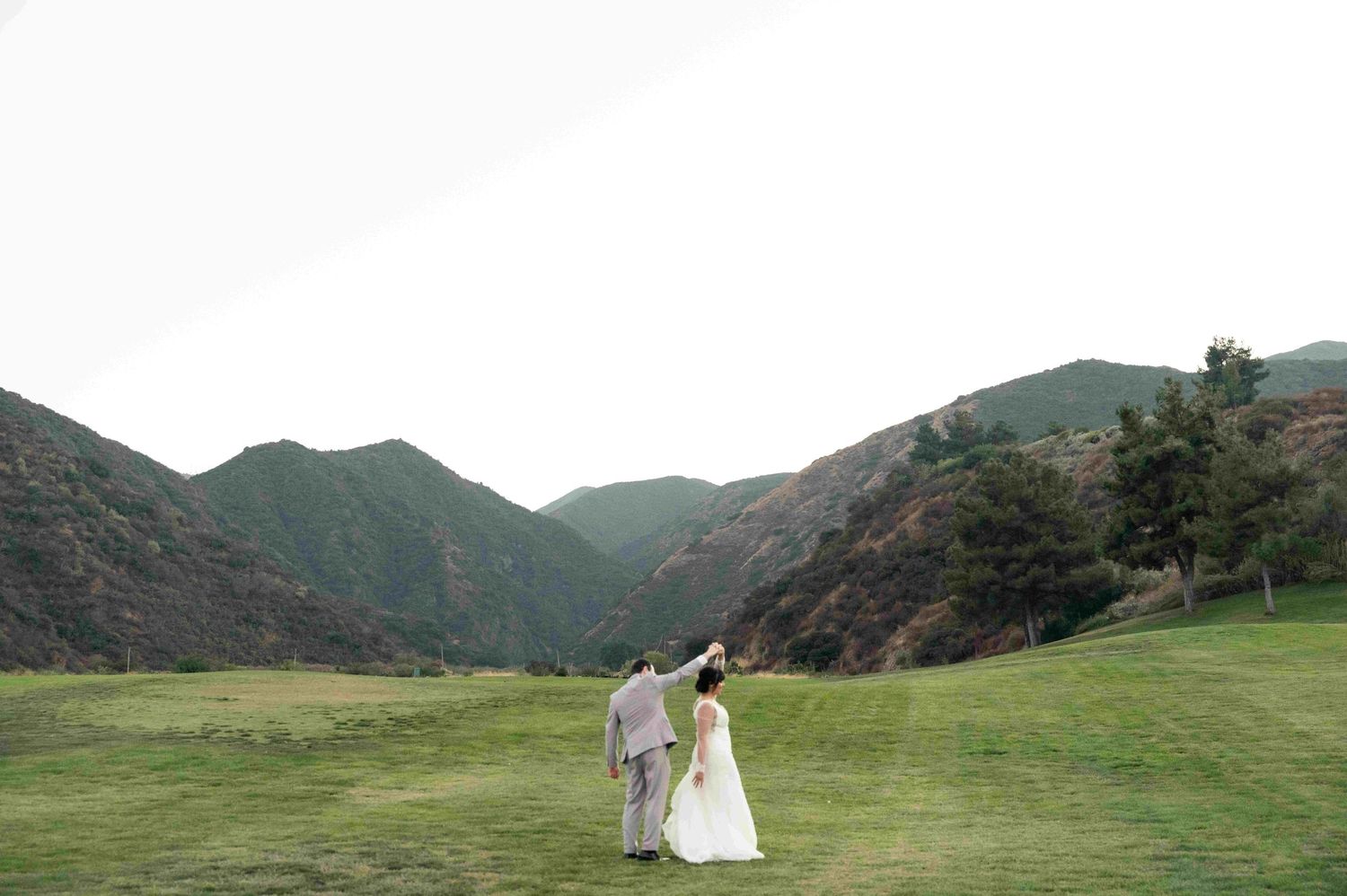 Newlyweds walk hand in hand across rolling green hills with dramatic mountain peaks in the background.