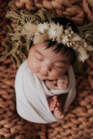 A sleeping newborn baby wearing white and adorned with delicate white flowers lies on a brown textured surface.