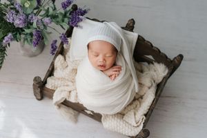 Newborn baby wrapped in white swaddle blanket lying in rustic wooden bowl with purple flowers nearby.