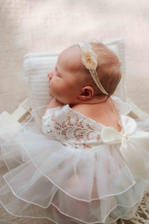 Sleeping newborn in delicate white lace dress with ruffled layers and headband lying on soft fabric.