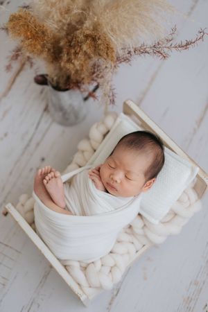 Peaceful newborn wrapped in white swaddle blanket resting in small wooden bed with dried floral decor.