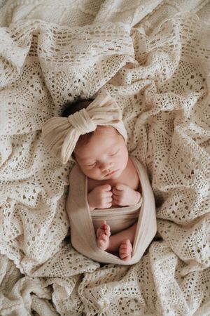 Newborn wrapped in soft beige fabric with matching bow headband rests peacefully on cream lace blanket.