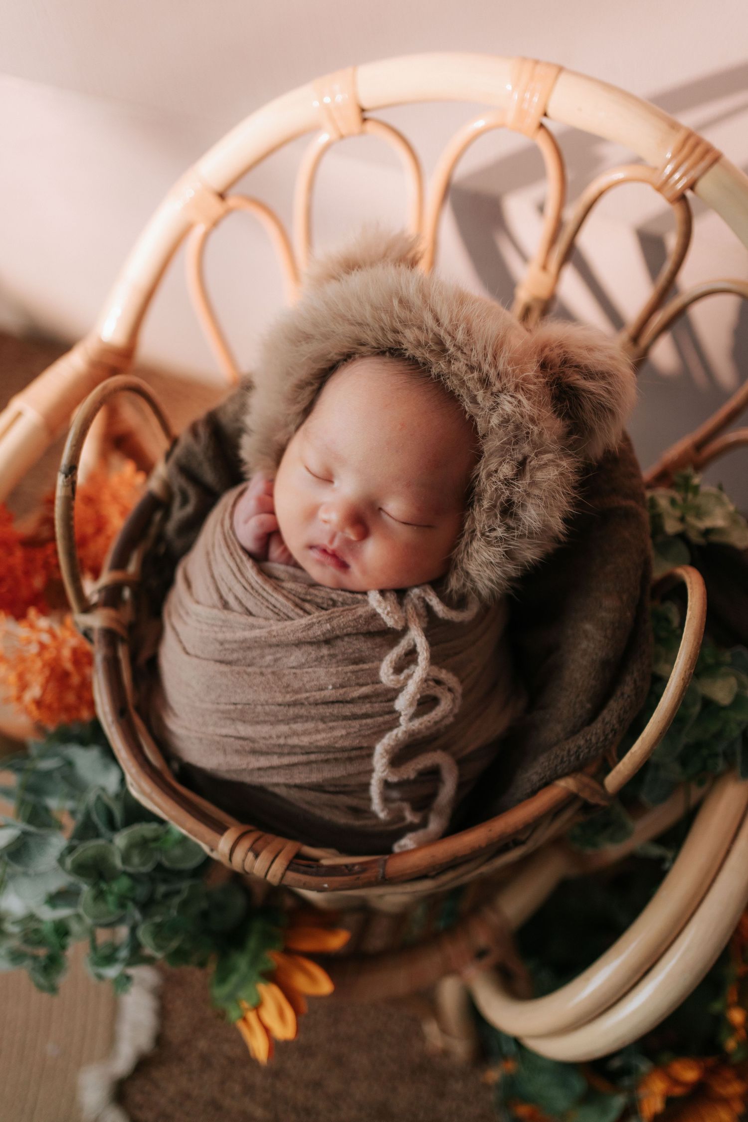 A sleeping newborn wrapped in brown swaddle blanket rests in a curved rattan chair with autumn decor.