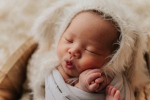 Peaceful sleeping baby nestled in soft white blankets with thumb in mouth during professional newborn photoshoot.