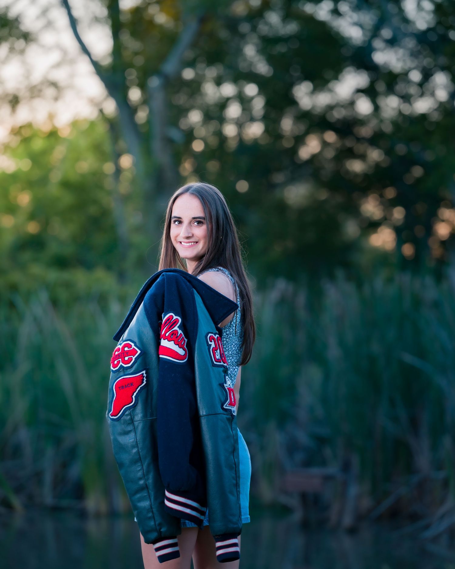 A smiling teenage girl poses outdoors in a varsity letterman jacket near a serene pond with lush green trees in the background.