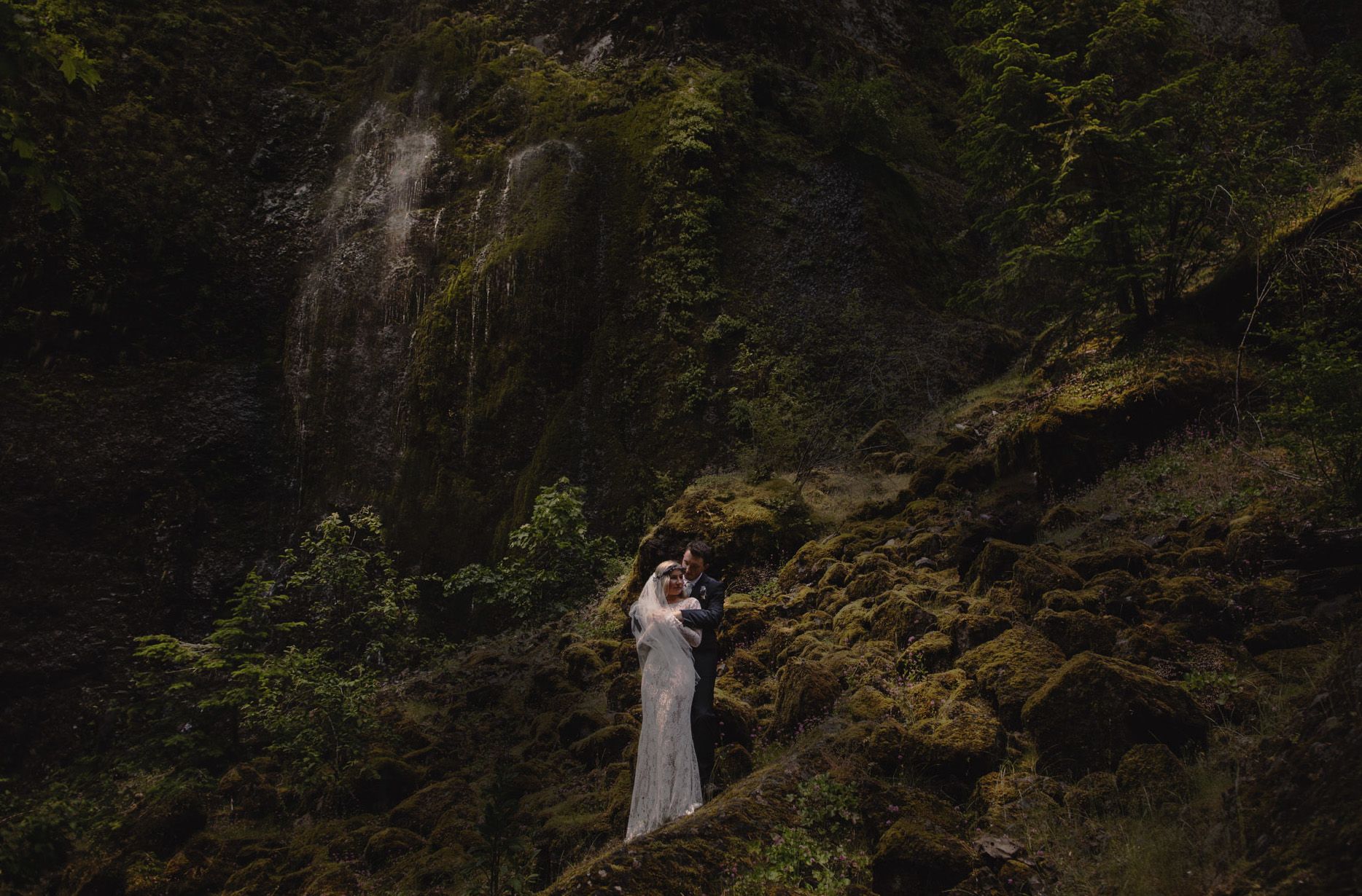 A couple in wedding attire embraces on mossy rocks before a lush waterfall in a dramatic forest setting.