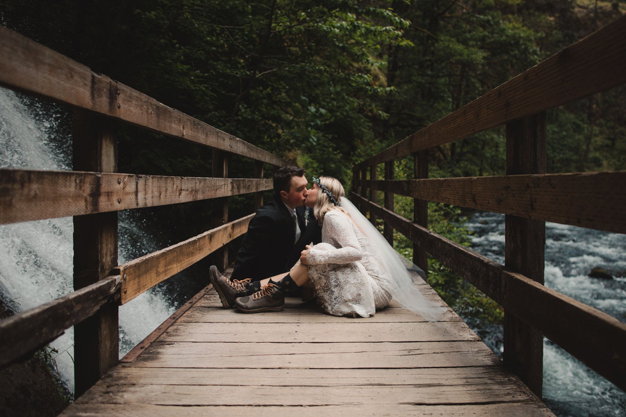 Bride and groom share a romantic kiss while sitting on a rustic wooden bridge beside a flowing river in a forest.