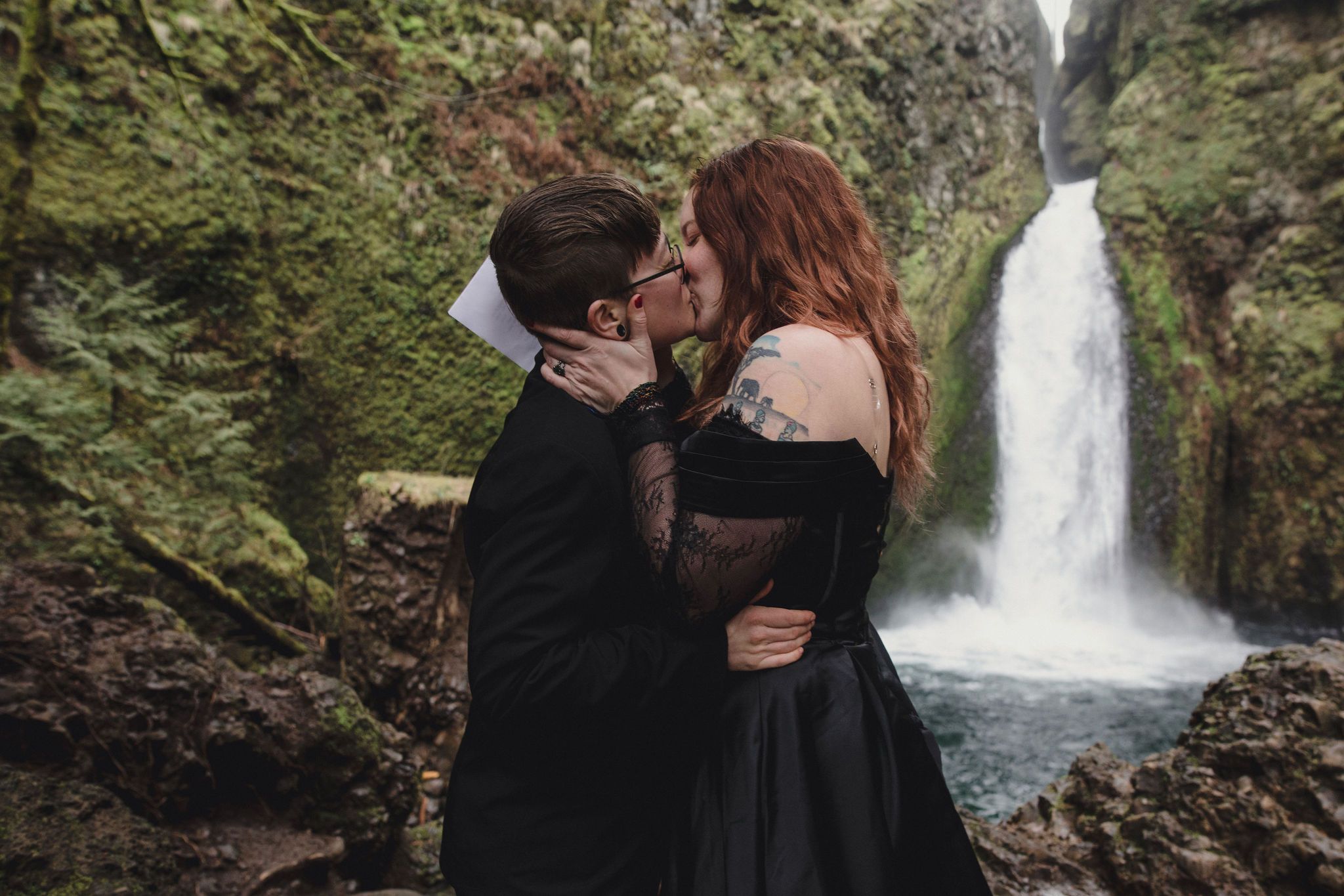 Couple in black wedding attire sharing a kiss in front of a lush green waterfall backdrop.