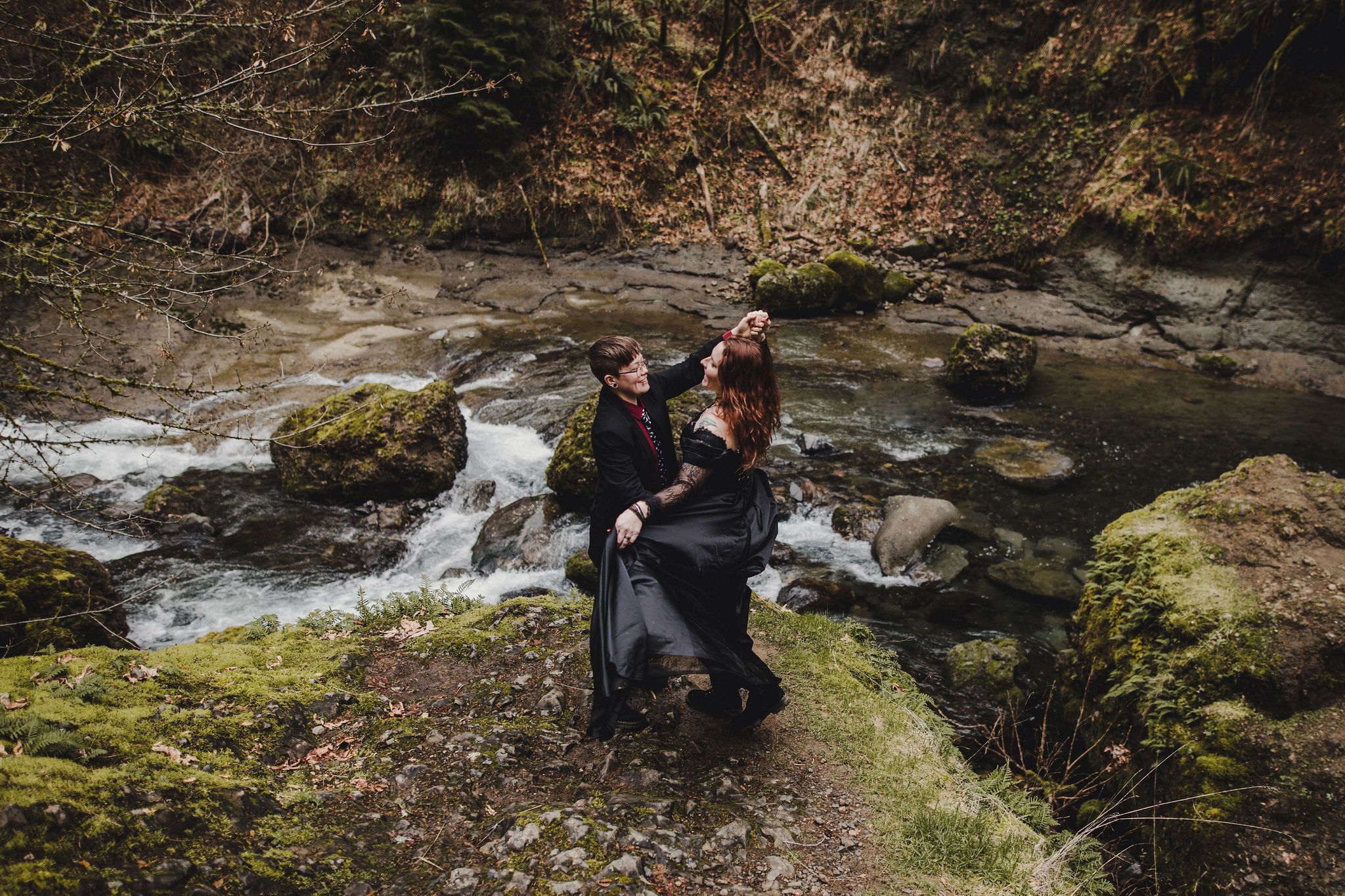 Couple embracing on mossy rocks beside a rushing forest stream surrounded by autumn trees.