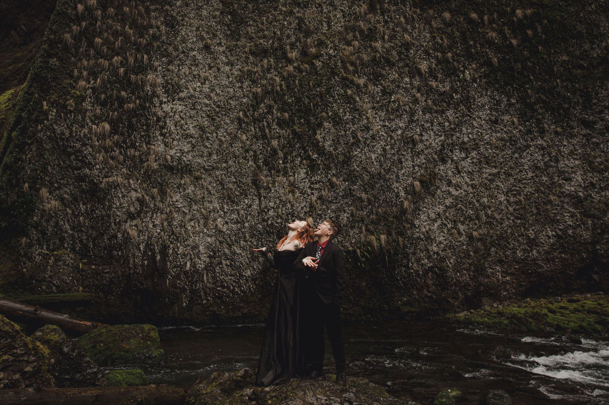 Couple in black attire embracing romantically against a mossy rock wall near a stream in a lush forest setting.