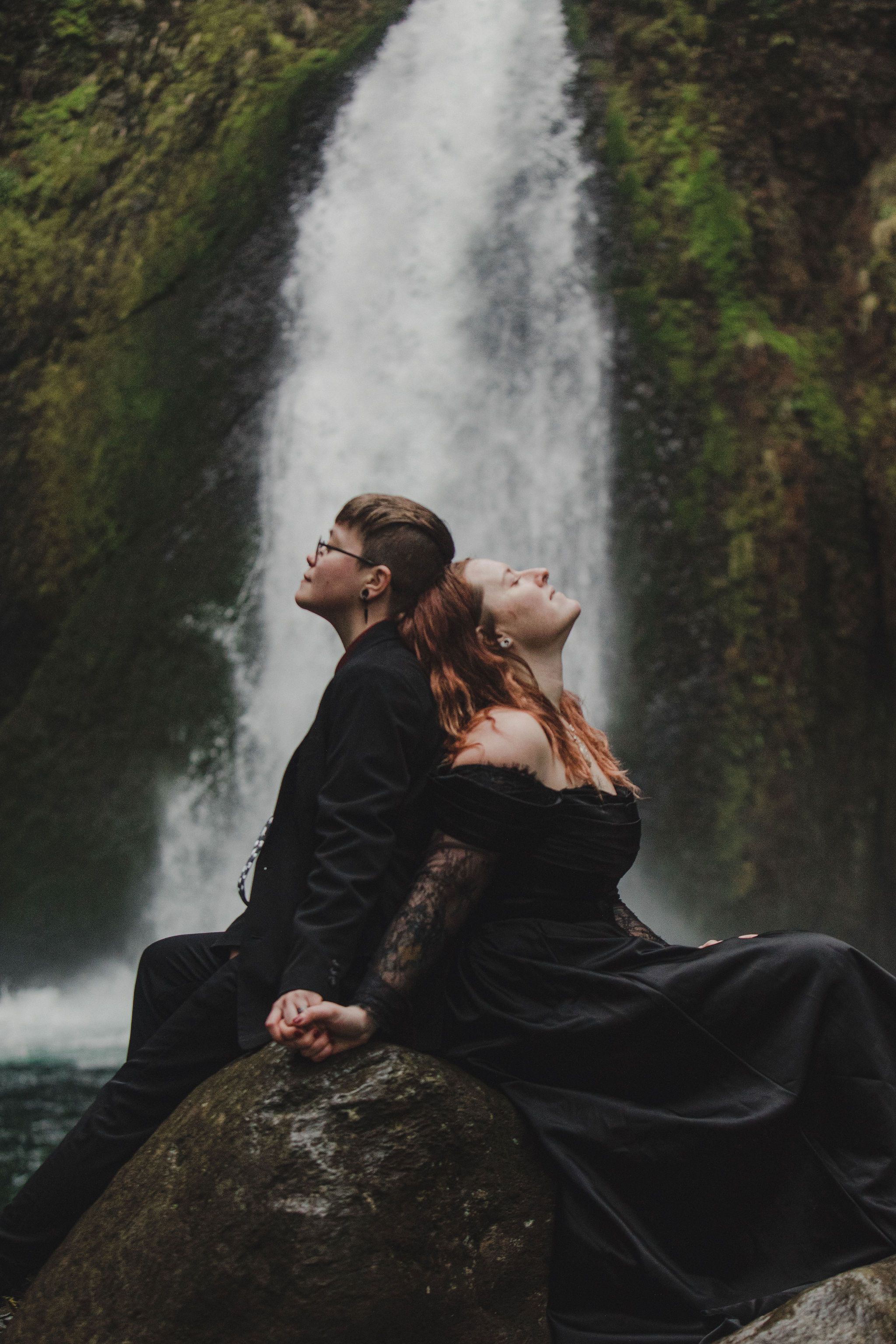 Couple in black gothic attire sitting back-to-back on a rock before a lush waterfall, holding hands.