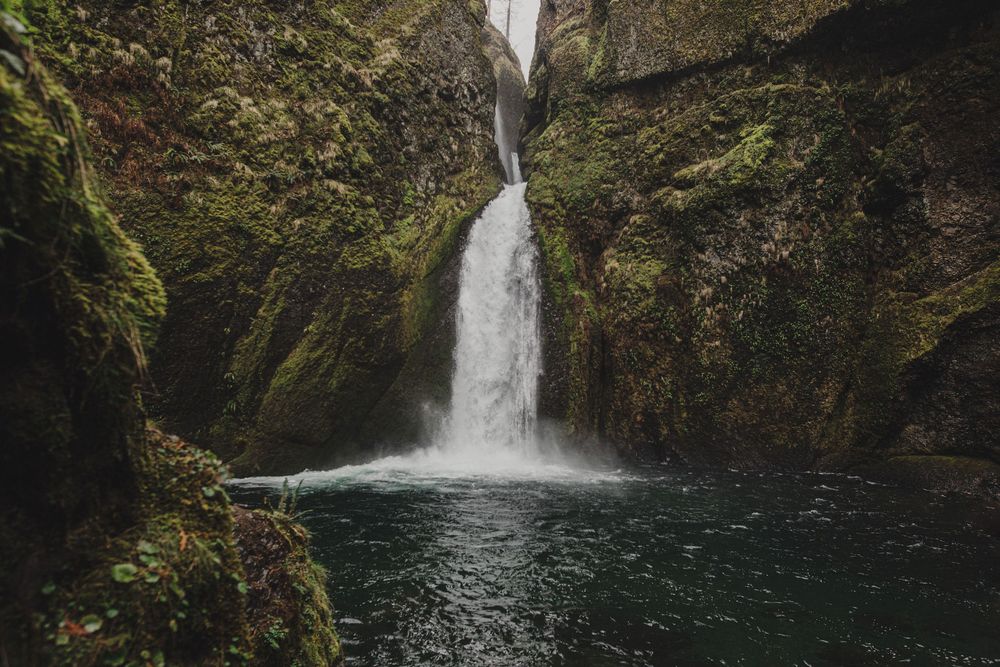 A stunning waterfall cascades into a serene pool surrounded by moss-covered rocky cliffs in a lush green gorge.