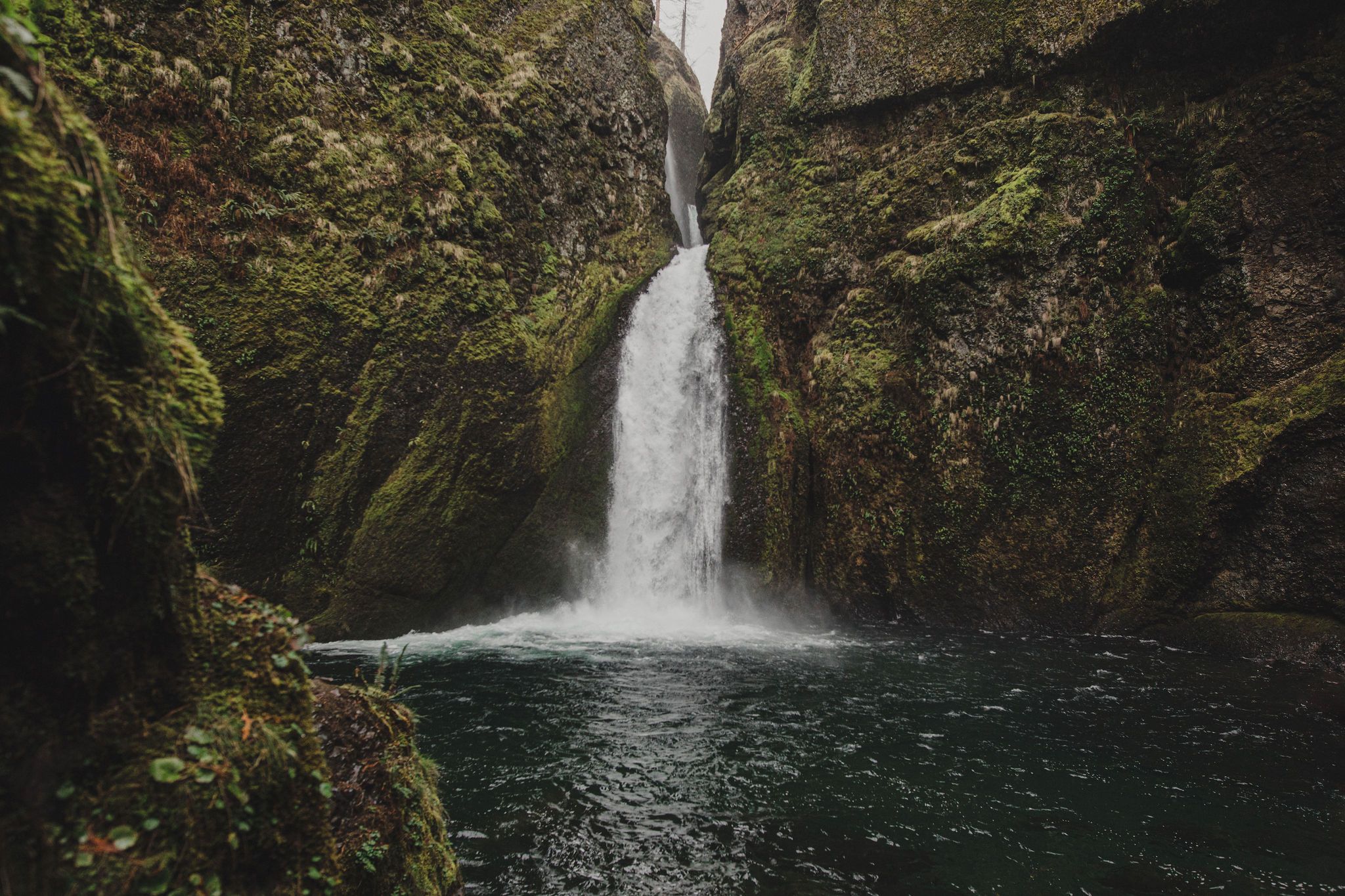 A stunning waterfall cascades into a serene pool surrounded by moss-covered rocky cliffs in a lush green gorge.