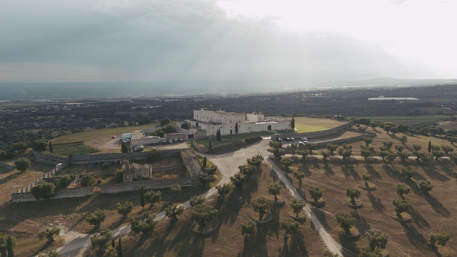 Aerial view of a large estate or winery surrounded by agricultural fields and olive trees under a cloudy sky.