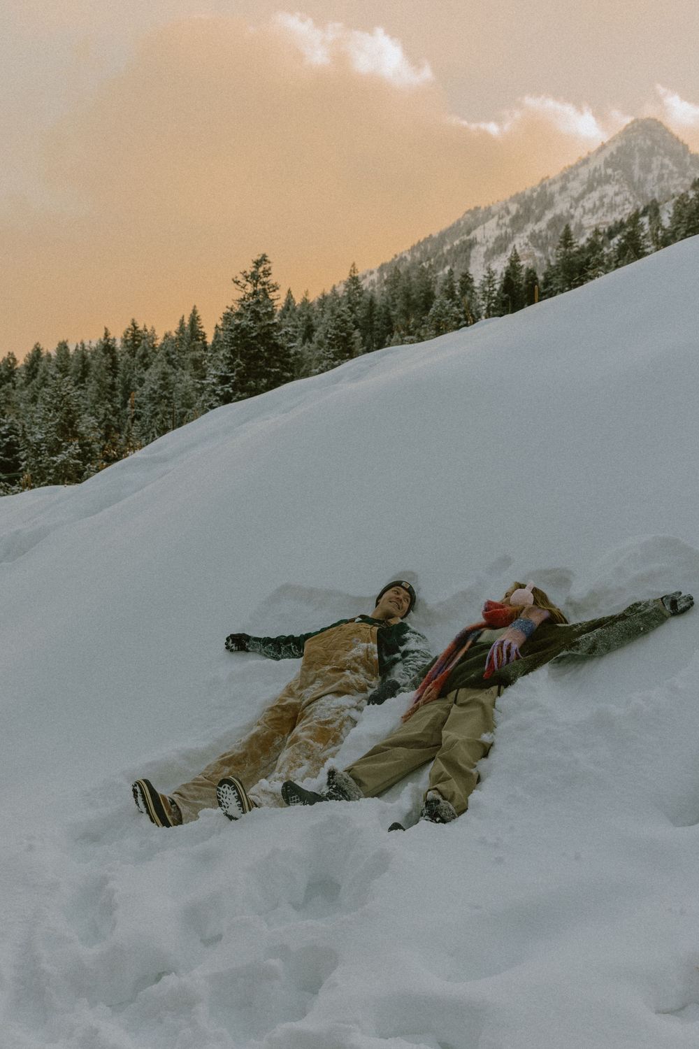 Two people lying in snow making snow angels on a snowy mountain slope at sunset with pine trees.