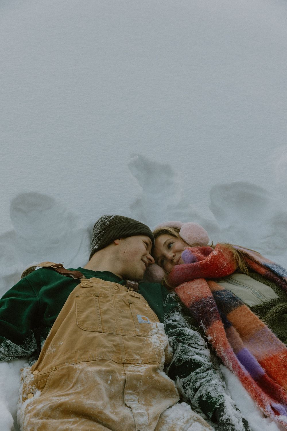 Couple lying in snow touching foreheads romantically, wearing winter clothes including overalls and colorful scarf.
