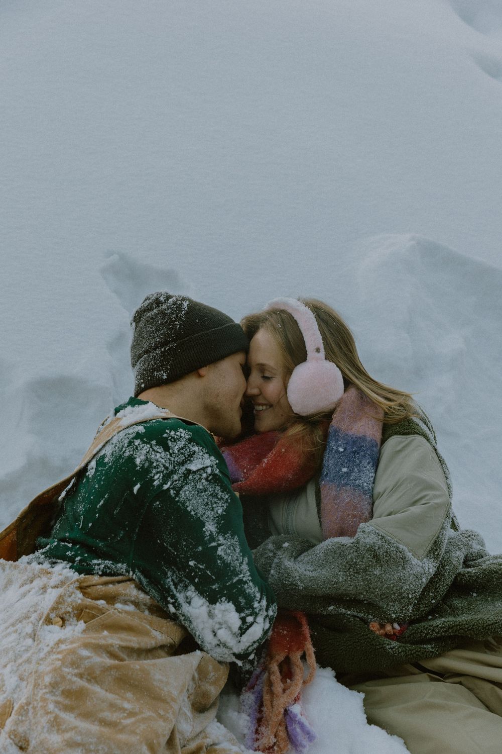 Couple embracing and kissing in snowy setting, woman wearing pink earmuffs and colorful scarf, wrapped in blanket.