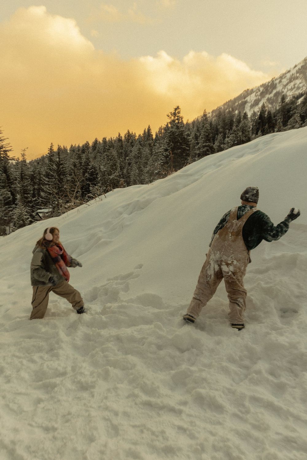 Two snowboarders play in deep snow on a mountain slope at golden hour sunset.