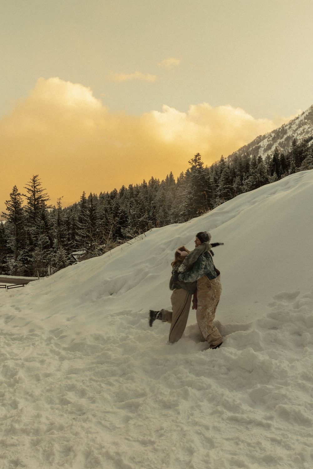 Two people embracing on a snowy mountain slope at sunset with snow-covered pine trees in the background.