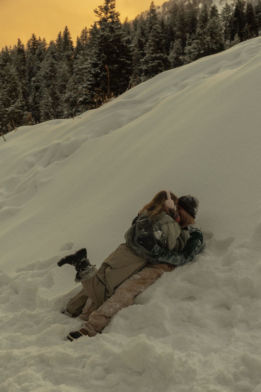 Couple embracing and lying together on a snowy hillside at sunset with pine trees in the background.