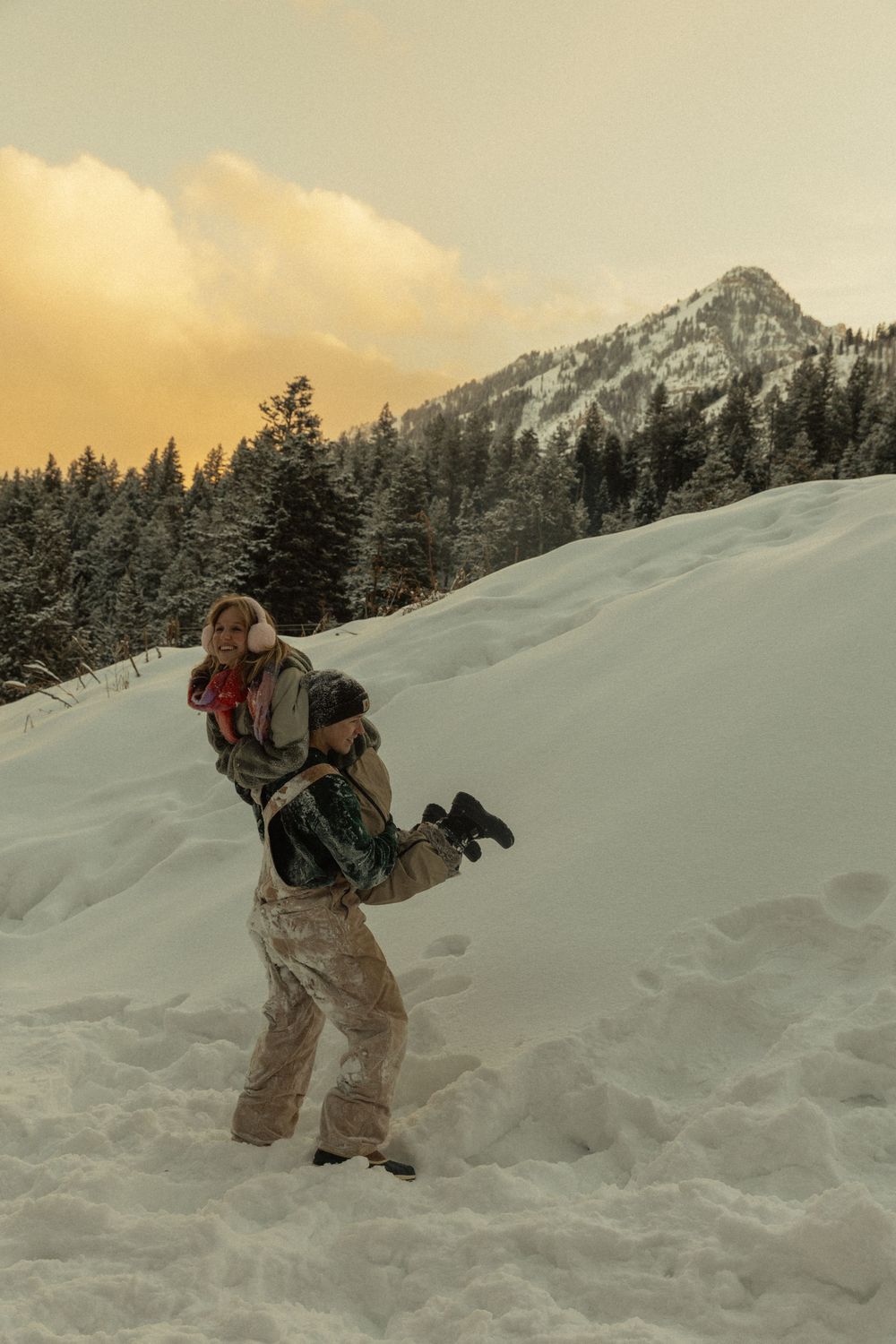 Couple playing in snow with snowy mountain and pine trees at sunset.