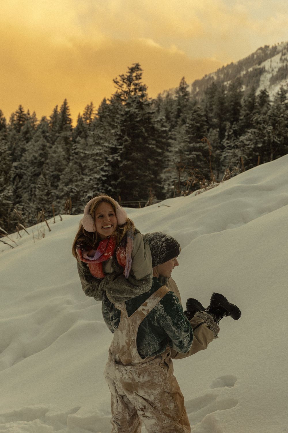 Couple playing in snow at sunset, man lifting woman, snowy forest and golden sky backdrop.