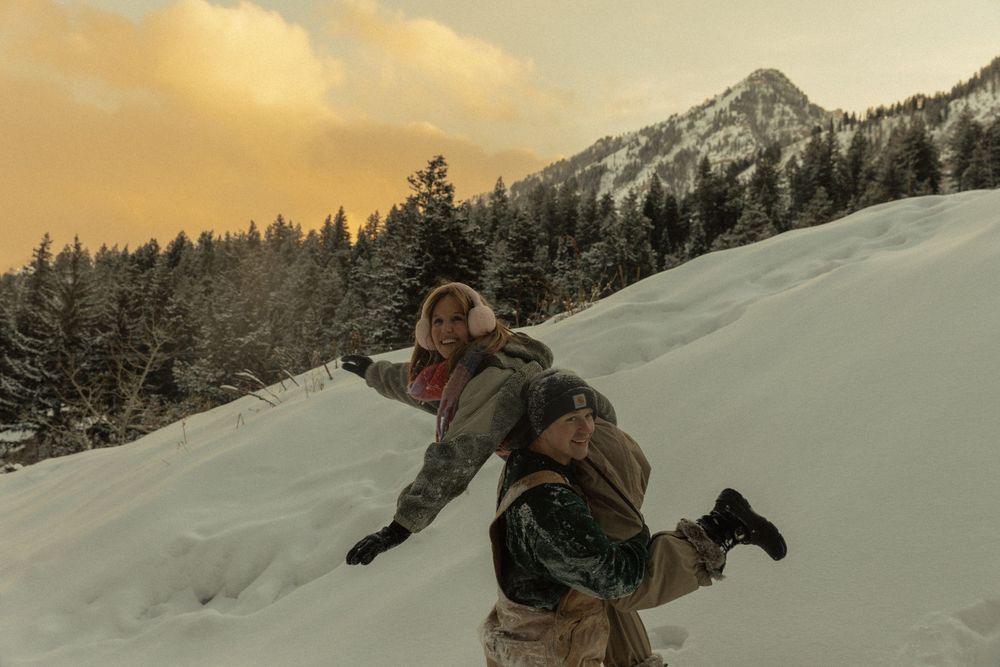Two people playing in snowy mountain landscape at sunset with pine trees and peaks in background.
