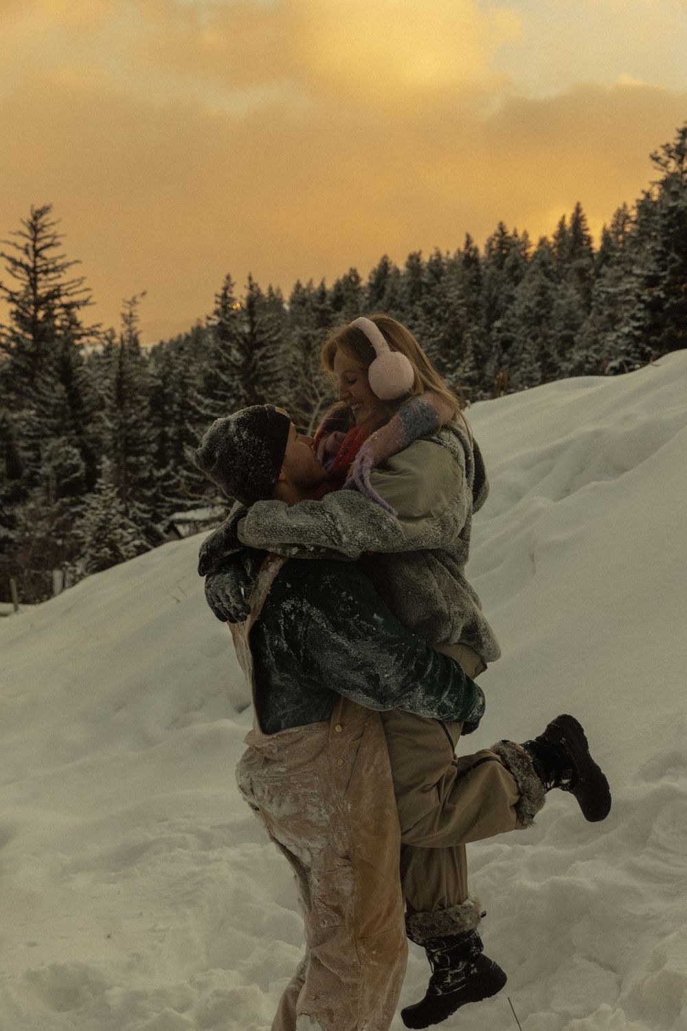 Couple sharing romantic kiss in snowy winter landscape at golden sunset with snow-covered pine trees.