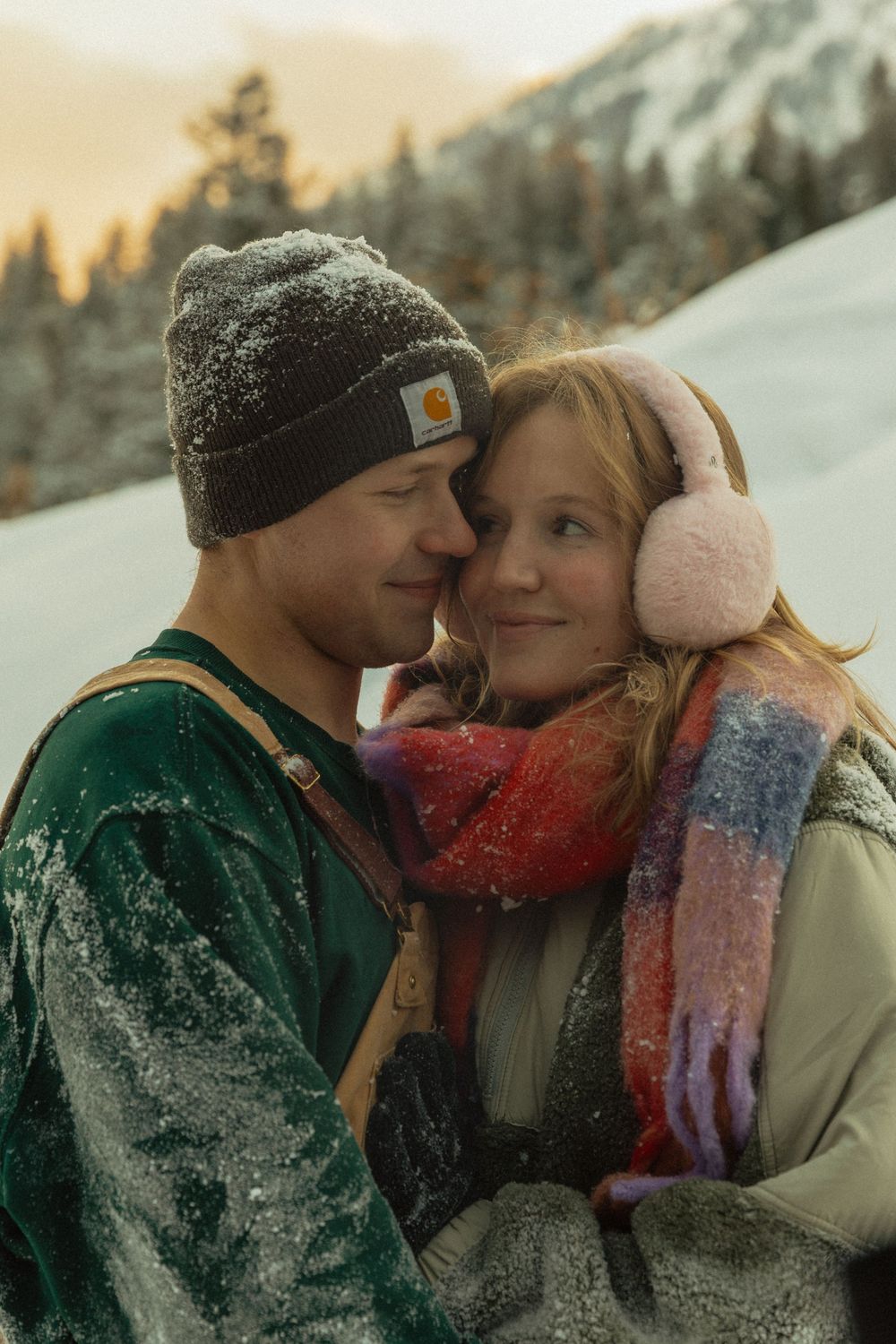 Couple in winter clothing snuggling close together in a snowy mountain landscape at golden hour.