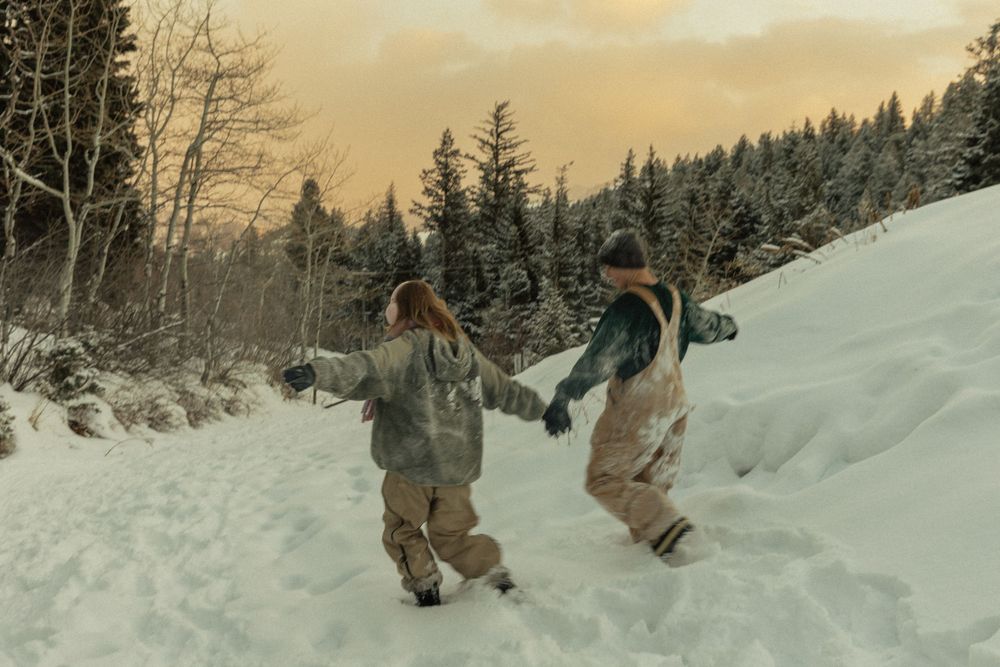 Two snowboarders hold hands while hiking up a snowy mountain trail surrounded by pine trees at sunset.