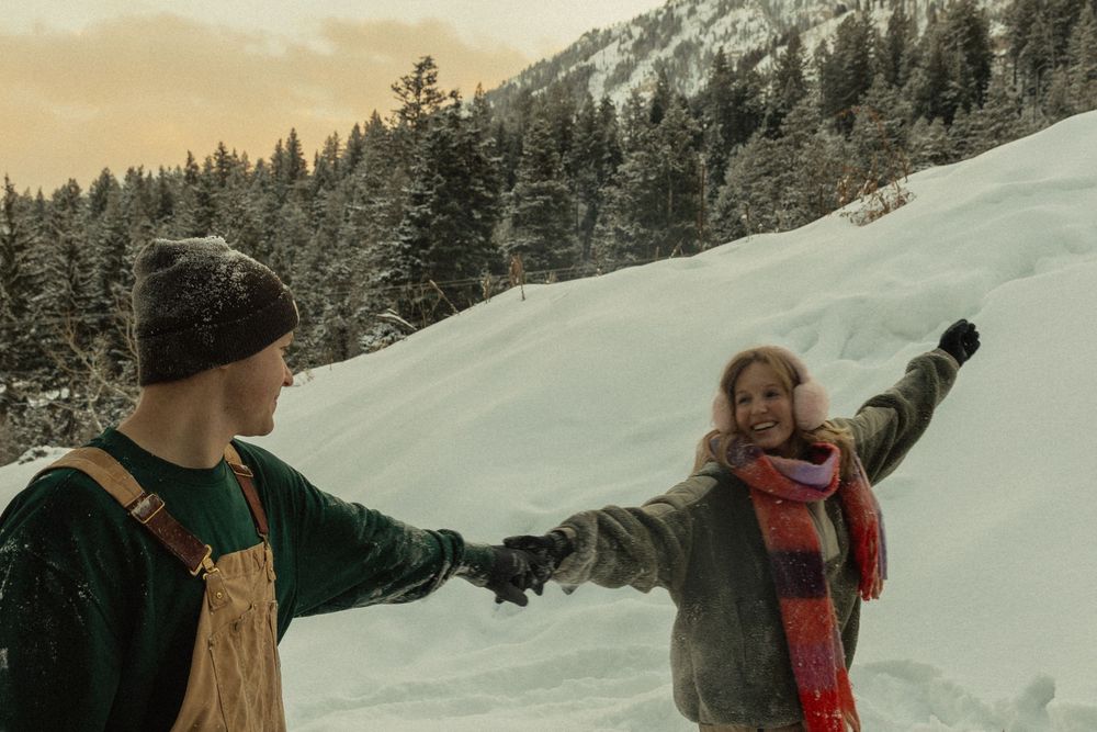 Couple holding hands in snowy winter landscape with snow-covered trees and mountains at sunset.