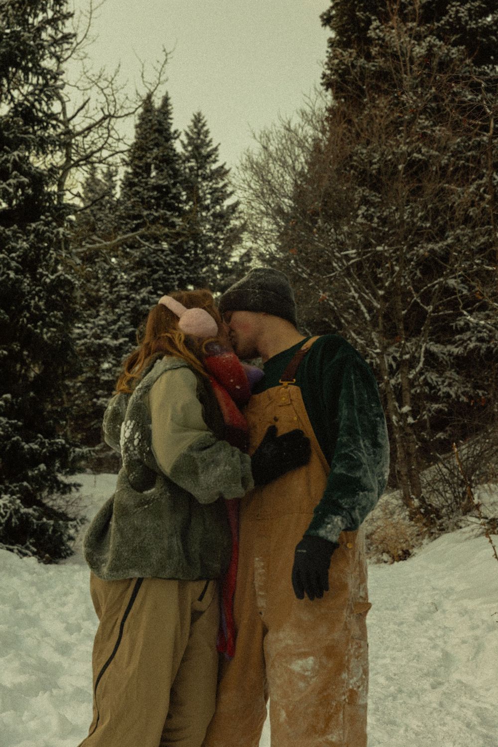 Couple sharing a romantic kiss in a snowy winter forest, wearing cozy layered outfits and gloves.
