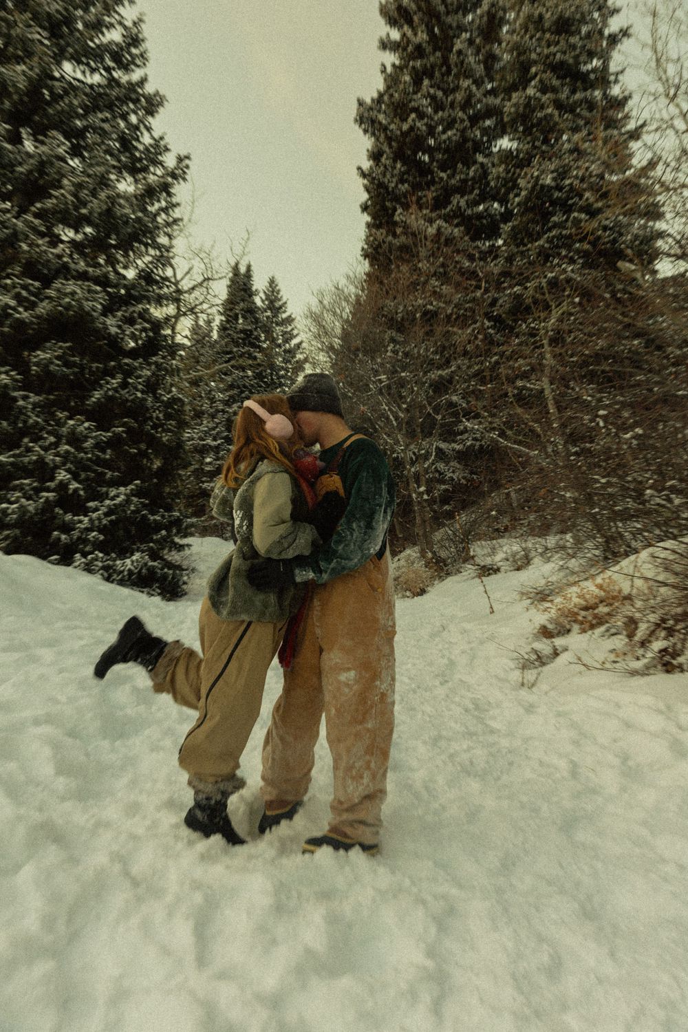 Couple embracing and kissing in a snowy winter forest surrounded by snow-covered evergreen trees.