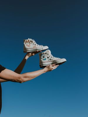 Hands hold two customized white high-top Converse Chuck Taylor sneakers against bright blue sky.