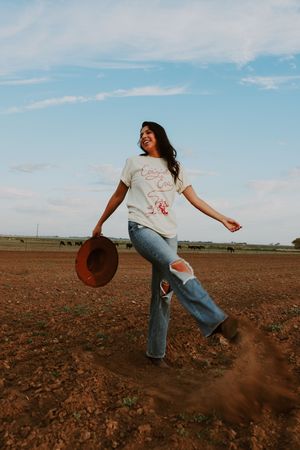 Joyful woman in Cowgirl Era tee and ripped jeans kicking up dirt on a farm field.
