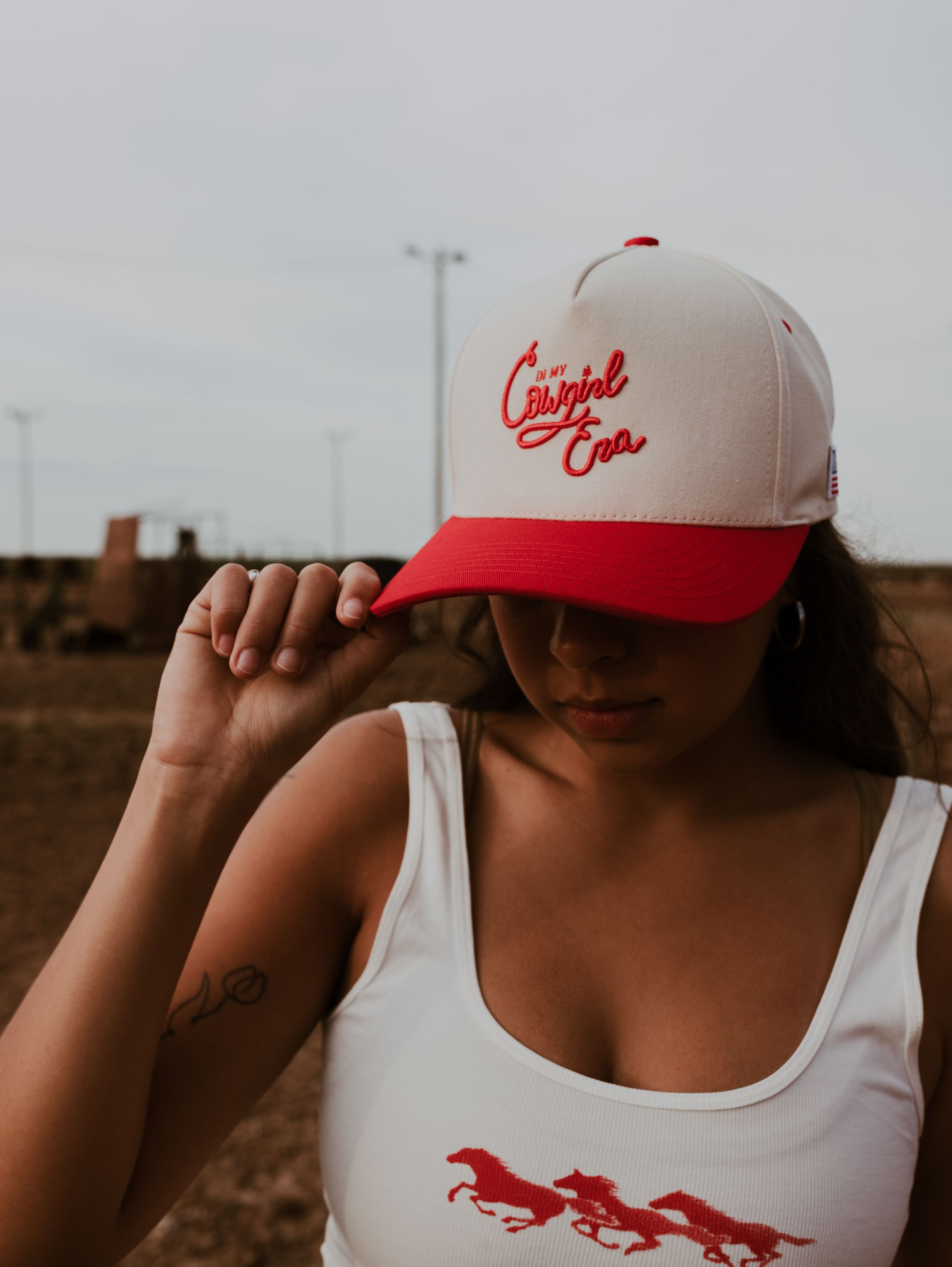 Woman wearing a white tank top and red Cowgirl Era trucker hat posing outdoors.