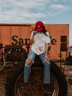 Woman in ripped jeans and red Cowgirl Era cap sitting on tractor near Santa Fe train car.