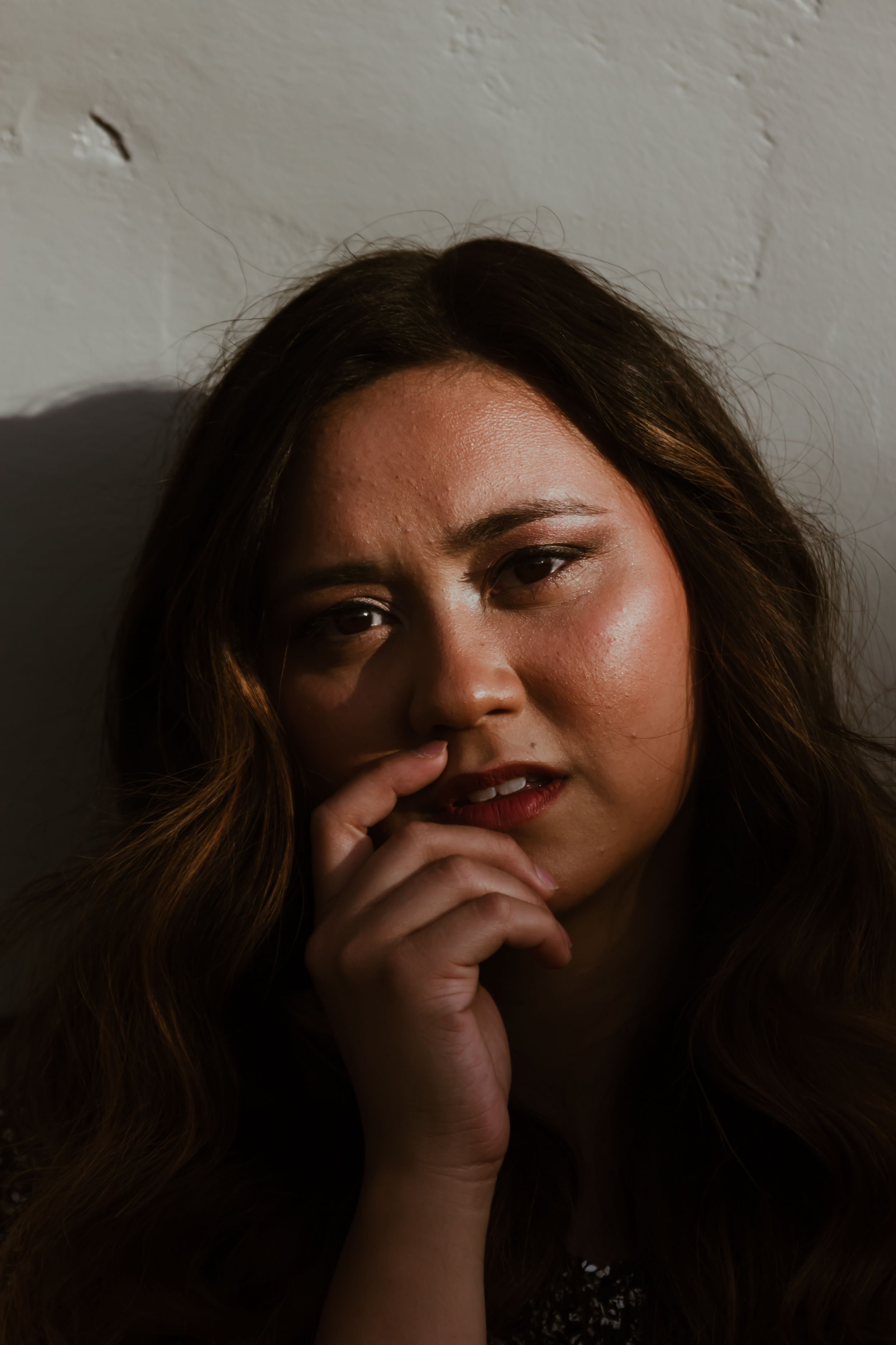Close-up portrait of brunette woman with freckles resting chin on hand against white textured wall.