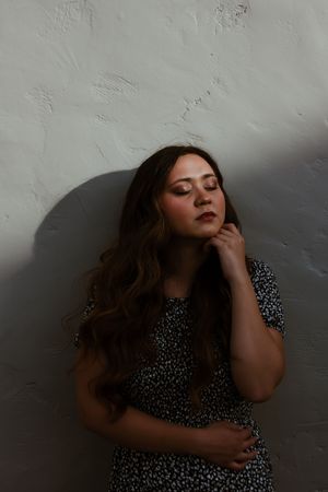 Brunette woman in floral dress leans against white textured wall with eyes closed in soft light.