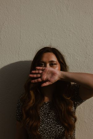 Brunette woman with wavy hair raises hand toward camera against textured beige wall in dramatic light.