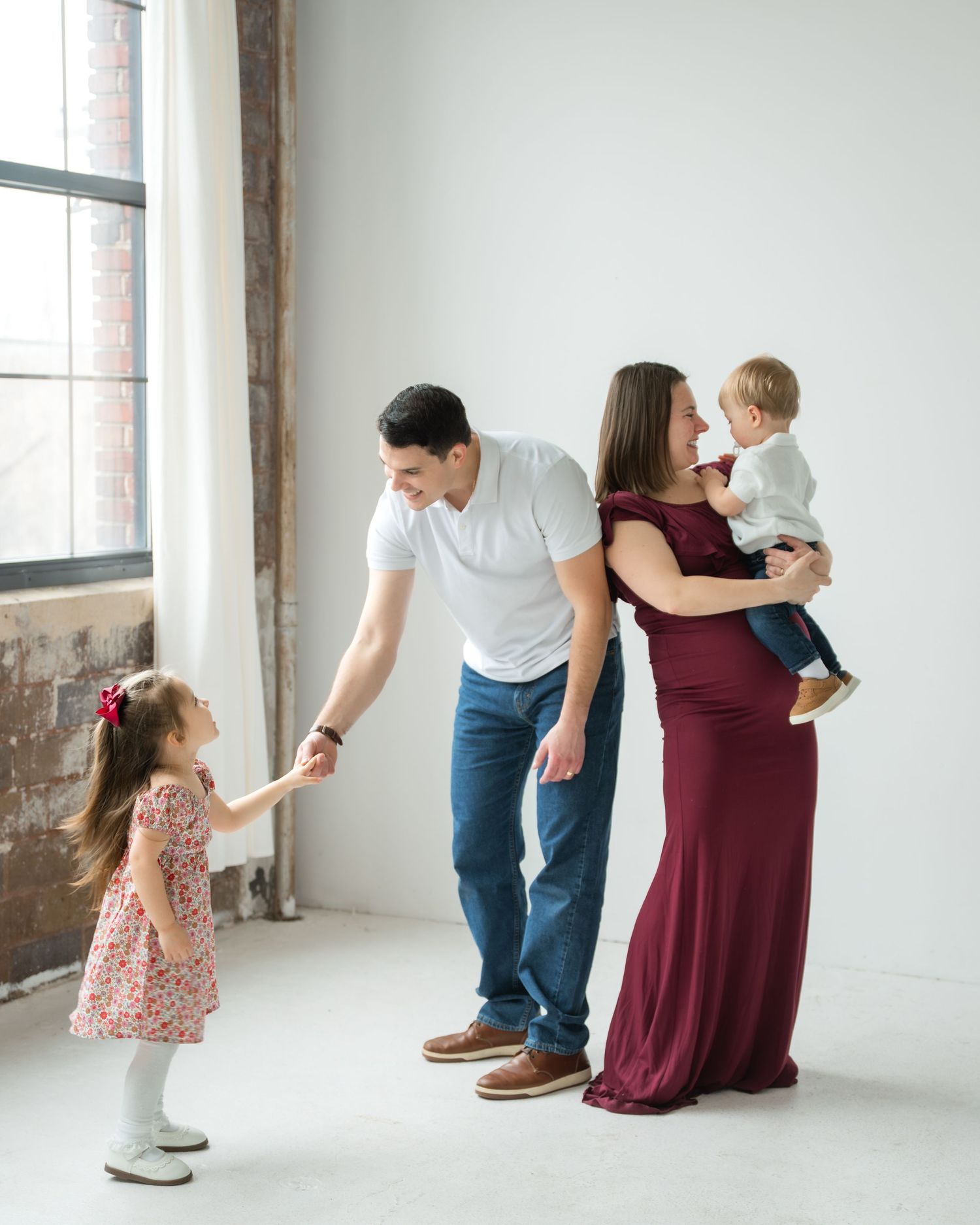 Family of four posing in bright studio, parents holding children, woman in burgundy gown, man in white shirt and jeans.
