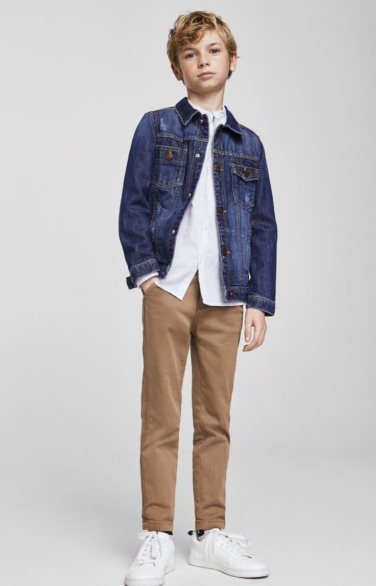 Boy wearing a dark blue denim jacket over a white shirt with khaki pants and white sneakers.