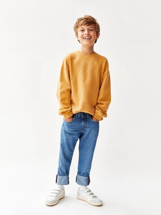 Smiling boy wearing a yellow sweatshirt, blue jeans, and white sneakers, posing against a white background.